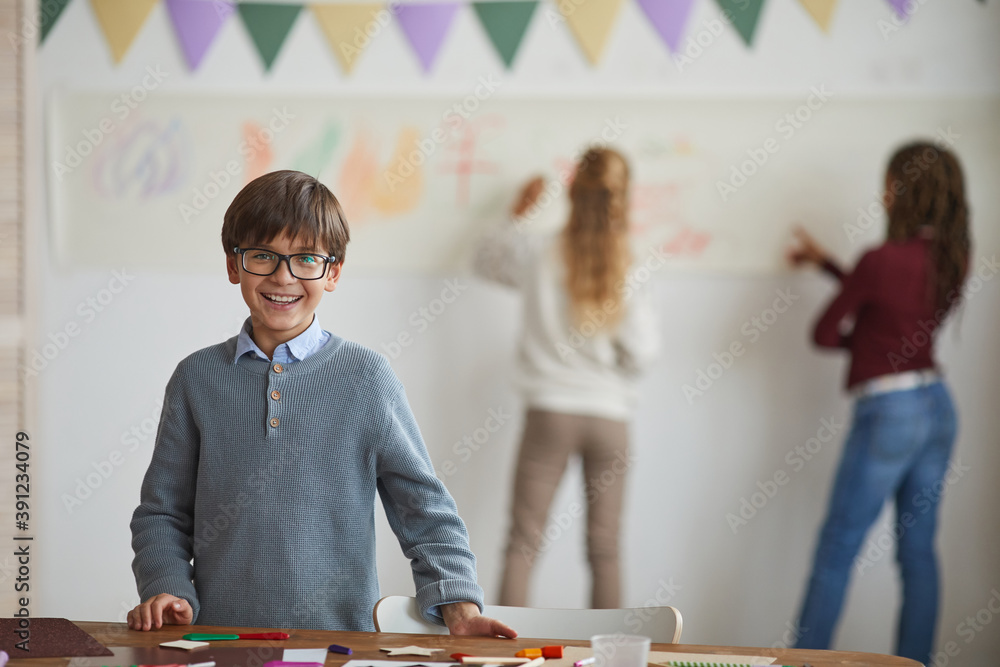 Portrait of smiling little boy wearing glasses and looking at camera while standing by crafting table and enjoying art class in school, copy space Portrait of smiling little boy wearing glasses and looking at camera while standing by crafting table and enjoying art class in school, copy space