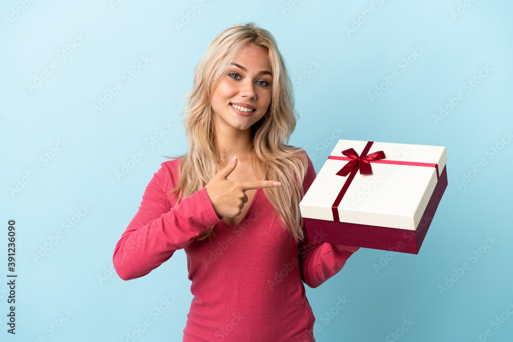 Young Russian woman holding a gift isolated on blue background and pointing it Young Russian woman holding a gift isolated on blue background and pointing it