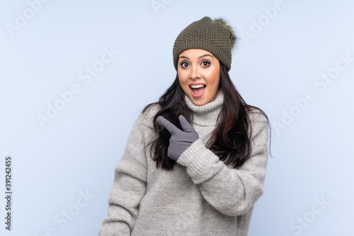 Young Colombian girl with winter hat over isolated blue background pointing finger to the side
