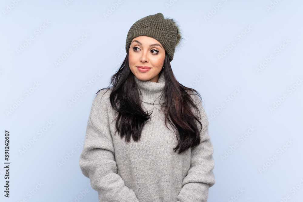 Young Colombian girl with winter hat over isolated blue background standing and looking to the side