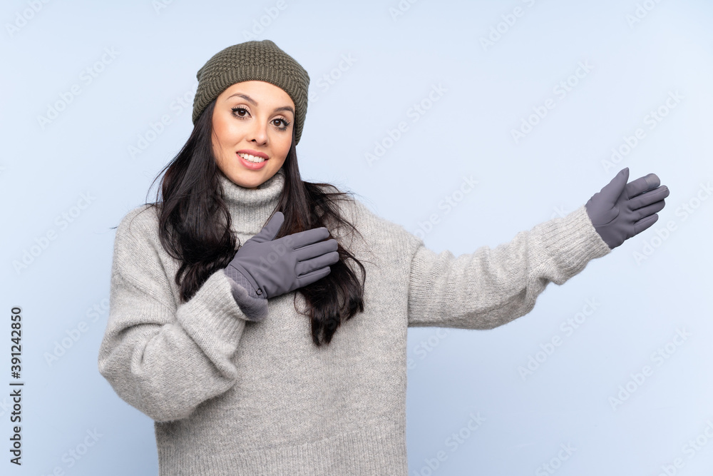 Young Colombian girl with winter hat over isolated blue background extending hands to the side for inviting to come Young Colombian girl with winter hat over isolated blue background extending hands to the side for inviting to come