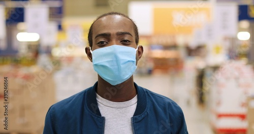 Close up portrait of happy young African American male in mask standing at supermarket indoors. Camera approaching handsome cheerful man in food store in quarantine. Virus concept