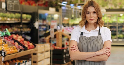 Happy young Caucasian woman manager in apron standing in supermarket indoors and looking at camera. Female beautiful worker at grocery store. Close up portrait concept. Retail business