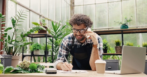 Happy Caucasian businessman talking on cellphone while standing in apron in small floral center and writing down order details. Joyful male florist calling on smartphone at work. Own business concept photo
