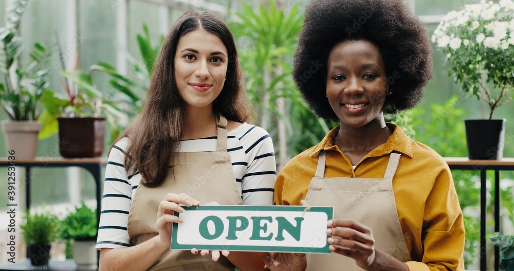 Close up portrait of joyful mixed-races female employees in aprons standing in floral shop holding Open sign in hands and smiling to camera. Caucasian and African American females opening flower store Close up portrait of joyful mixed-races female employees in aprons standing in floral shop holding Open sign in hands and smiling to camera. Caucasian and African American females opening flower store