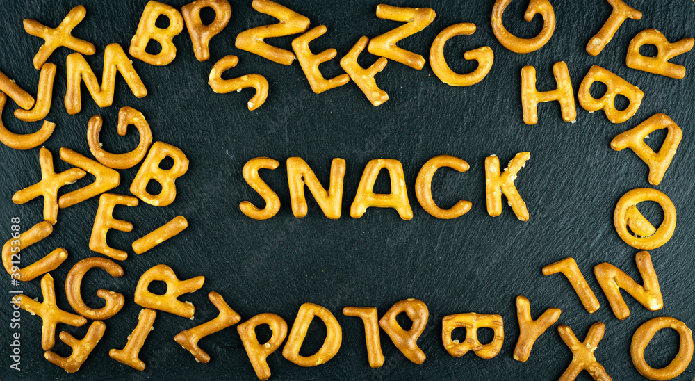 Snack text among salty sticks letters, alphabet on black stone background. Junk food, unhealthy diet concept.