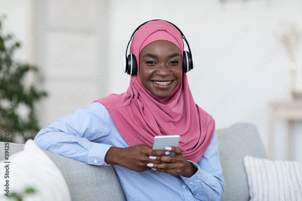 Cheerful Black Muslim Female Listening Music On Smartphone At Home, Wearing Headphones