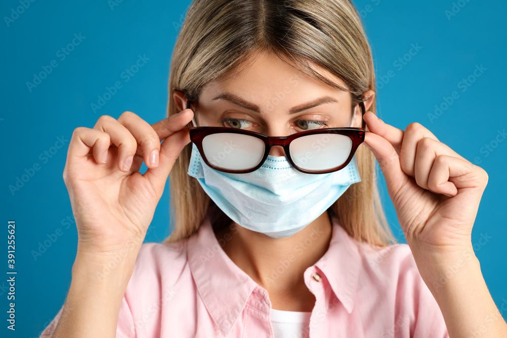 Woman with foggy glasses caused by wearing disposable mask on blue background. Protective measure during coronavirus pandemic Woman with foggy glasses caused by wearing disposable mask on blue background. Protective measure during coronavirus pandemic