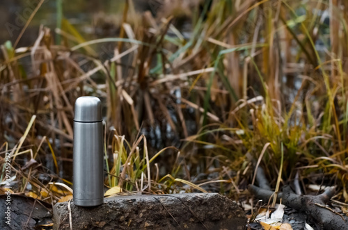 Vacuum steel thermos outdoors. Aluminum Travel Flask stands by the water. Hike concept, relaxation in nature, hot drink in cold weather.