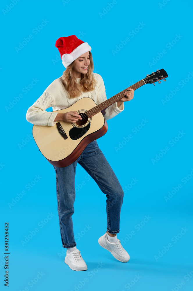 Young woman in Santa hat playing acoustic guitar on light blue background. Christmas music Young woman in Santa hat playing acoustic guitar on light blue background. Christmas music