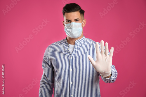Man in protective face mask and medical gloves showing stop gesture against pink background, focus on hand