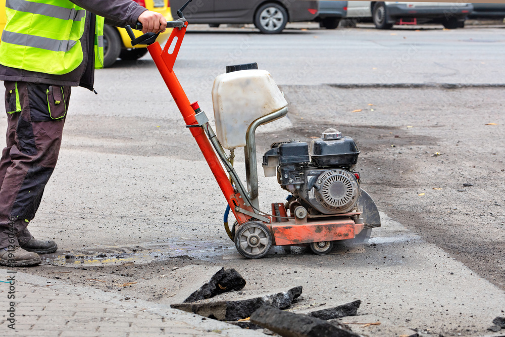 A road builder in reflective overalls uses a portable asphalt cutter to cut asphalt to repair a section of the roadway.
