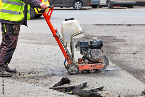 A road builder in reflective overalls uses a portable asphalt cutter to cut asphalt to repair a section of the roadway.