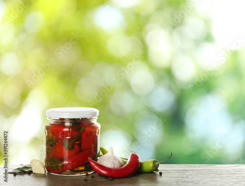 Jar of tasty pickled chili peppers on wooden table against blurred green background. Space for text
