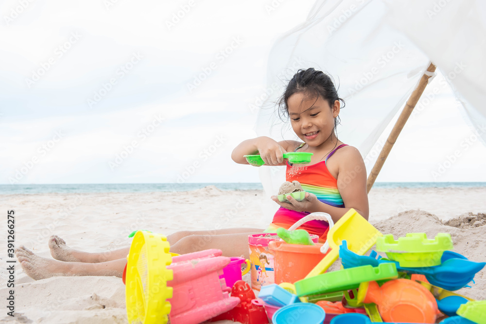 Fototapeta premium cute little girl playing on the beach on summer holidays. Happy child playing with sand at the beach. kid wear brightly colored swimwear and colorful toys.