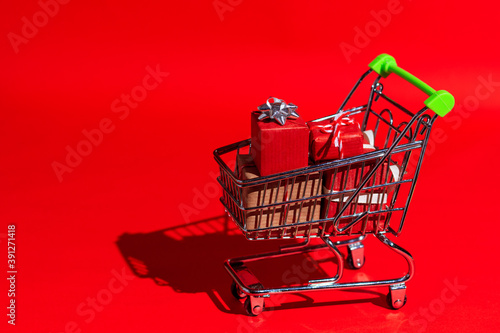 Supermarket trolley full of Christmas gifts on a red background in pop-art style with hard shadow. Gifts theme for new year, christmas, black friday.