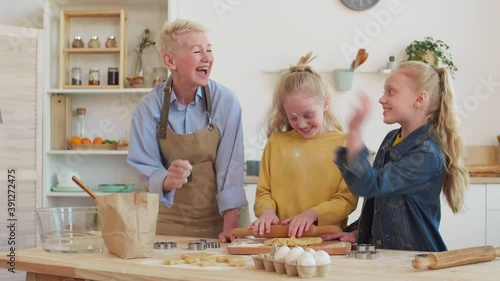 Medium of white-haired Caucasian grandma smearing with flour blue-eyed little twin granddaughters, girls and woman starting playing, throwing gough ingredient at each other, having fun, laughing photo