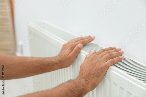 Man warming hands on heating radiator near white wall, closeup