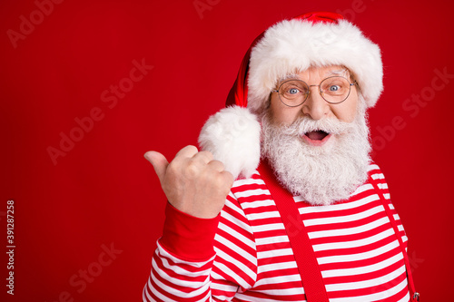 Closeup photo of retired grandpa white beard direct finger empty space suggest winter design wear santa x-mas costume suspenders spectacles striped shirt cap isolated red color background © deagreez