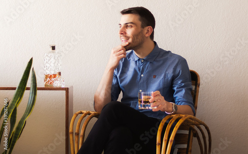 Photos Retrato de un hombre adulto caucásico y apuesto relajado sentado en un sillón de mimbre disfrutando de un vaso de whisky escocés en un ambiente cálido y confortable en su hogar