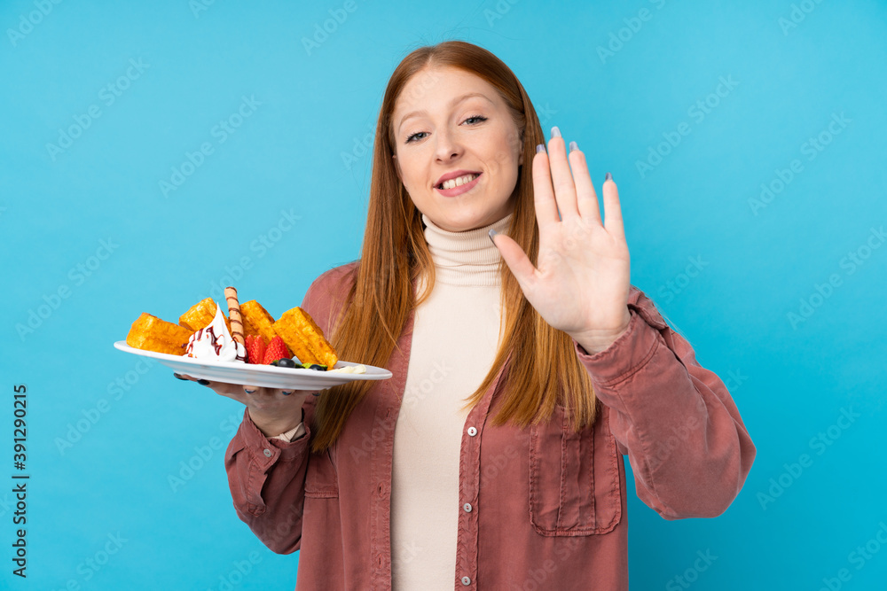 Young redhead woman holding waffles over isolated background saluting with hand with happy expression Young redhead woman holding waffles over isolated background saluting with hand with happy expression