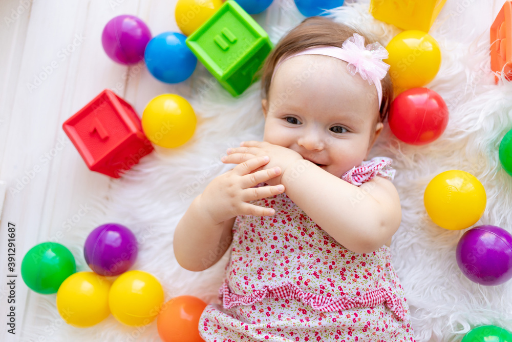 a small child girl lies in red clothes on a white Mat among toys balls and cubes and sucks her finger a small child girl lies in red clothes on a white Mat among toys balls and cubes and sucks her finger