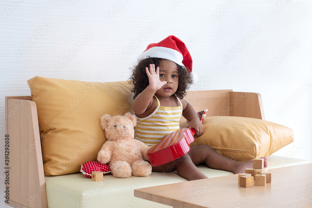 Obraz premium Cute dark skinned little girl wearing Santa hat and waving hand to someone on couch at home, new year, x-mas, Christmas concept