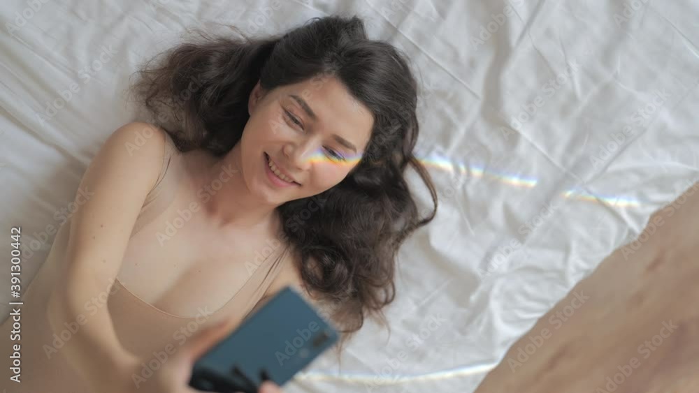 Happiness, lgbt, lesbian concept. A happy mixed race woman is taking selfie photo with rainbow flare on her face on her smartphone while lying on bed in sunny room. Happiness, lgbt, lesbian concept. A happy mixed race woman is taking selfie photo with rainbow flare on her face on her smartphone while lying on bed in sunny room.