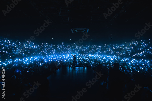 Vocalist in front of crowd on scene in 
stadium. Bright stage lighting, crowded dance floor. Phone lights at concert. Band blue silhouette crowd. People with cell phone lights.