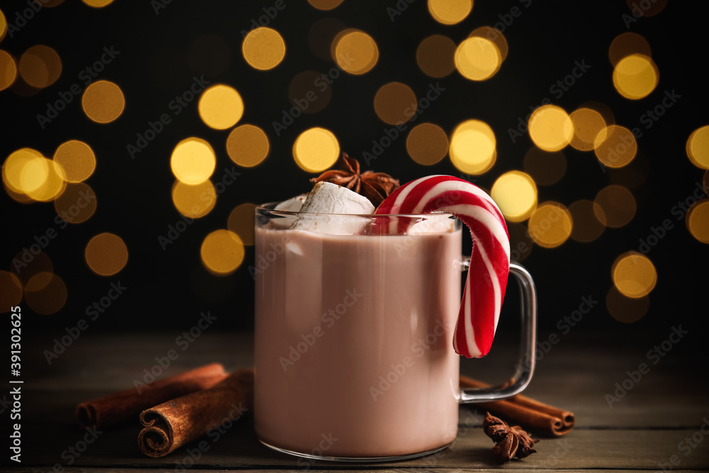 Obraz premium Glass cup of tasty cocoa with marshmallows and Christmas candy cane on wooden table against blurred festive lights