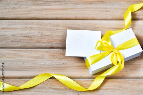 Christmas present with yellow ribbon and empty paper card on wooden background. Christmas gifts, seasonal holidays greetings concept. Flat lay, copy space.