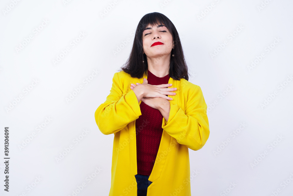 Caucasian woman standing against white background closes eyes and keeps hands on chest near heart, expresses sincere emotions, being kind hearted and honest. Body language and real feelings concept. Caucasian woman standing against white background closes eyes and keeps hands on chest near heart, expresses sincere emotions, being kind hearted and honest. Body language and real feelings concept.