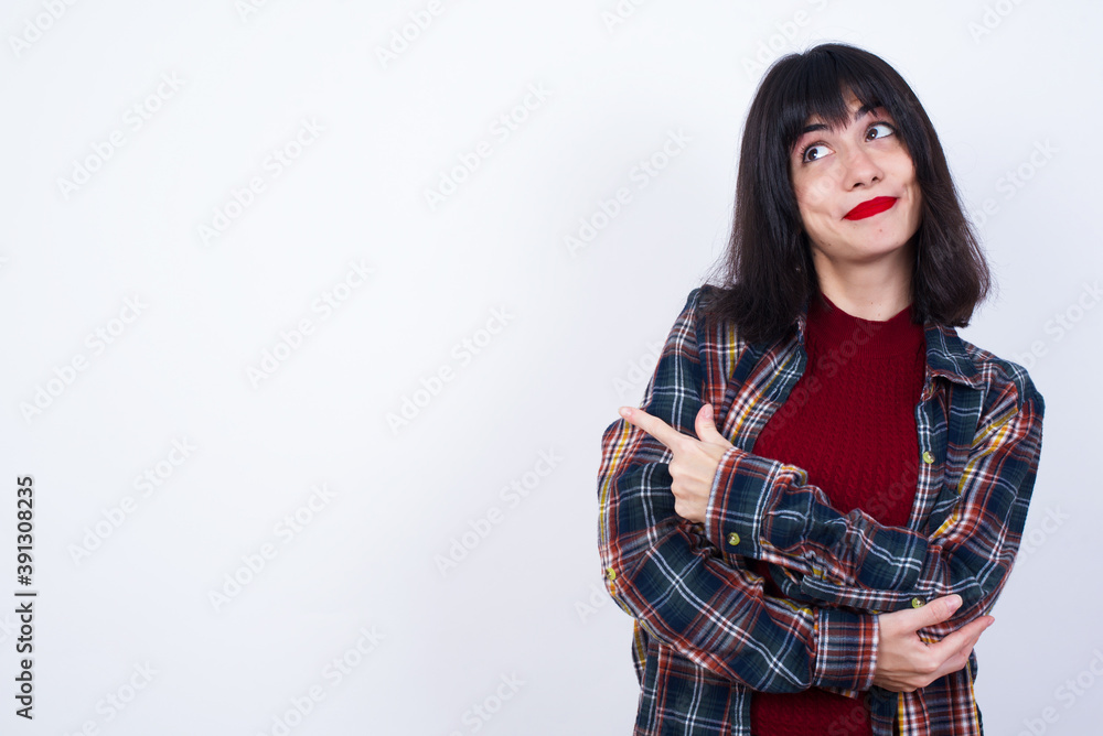 Portrait of Beautiful young Caucasian woman standing against white background posing on camera with tricky look, presenting product with index finger. Advertisement concept. Portrait of Beautiful young Caucasian woman standing against white background posing on camera with tricky look, presenting product with index finger. Advertisement concept.