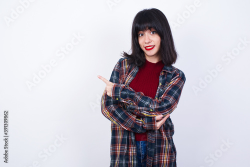 Beautiful young Caucasian woman standing against white background smiling broadly at camera, pointing fingers away, showing something interesting and exciting. © Jihan