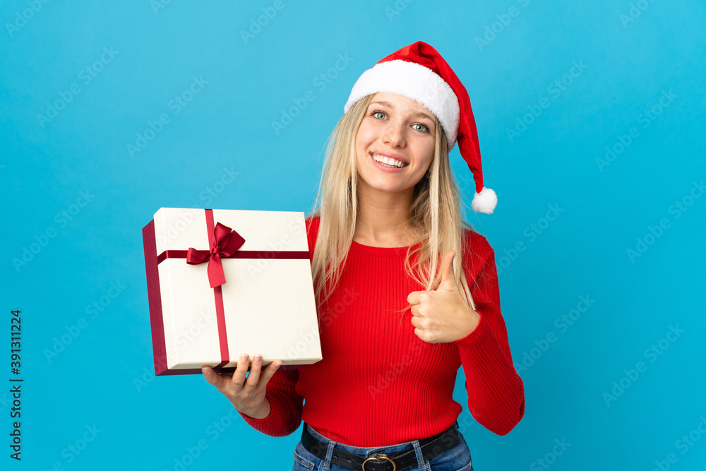 Woman with christmas hat holding a present isolated on blue background with thumbs up because something good has happened Woman with christmas hat holding a present isolated on blue background with thumbs up because something good has happened