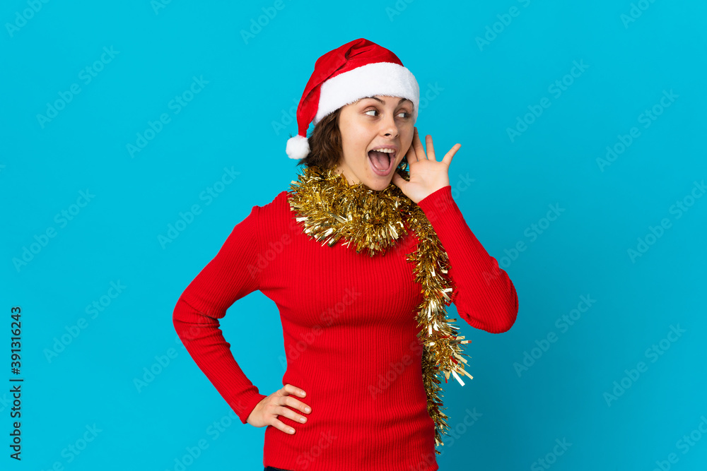 English girl with christmas hat isolated on blue background listening to something by putting hand on the ear English girl with christmas hat isolated on blue background listening to something by putting hand on the ear