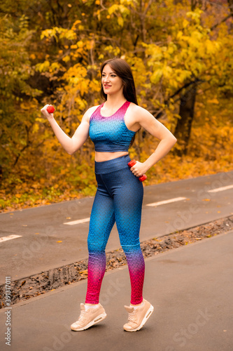 Fototapeta Naklejka Na Ścianę i Meble -  Women training in the park. Sports girl in a bright sportswear. Girl smiling and holding dumbbells in her arms.
