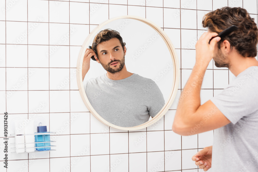 Young man looking at camera while combing hair on blurred foreground in bathroom Young man looking at camera while combing hair on blurred foreground in bathroom