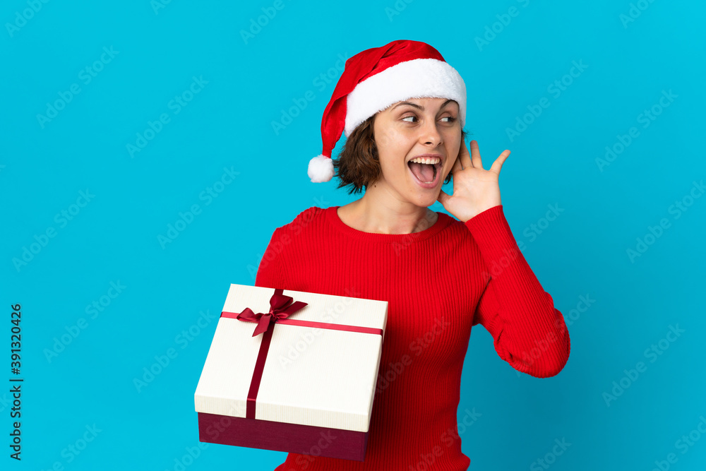 English girl with christmas hat holding a present isolated on blue background listening to something by putting hand on the ear English girl with christmas hat holding a present isolated on blue background listening to something by putting hand on the ear