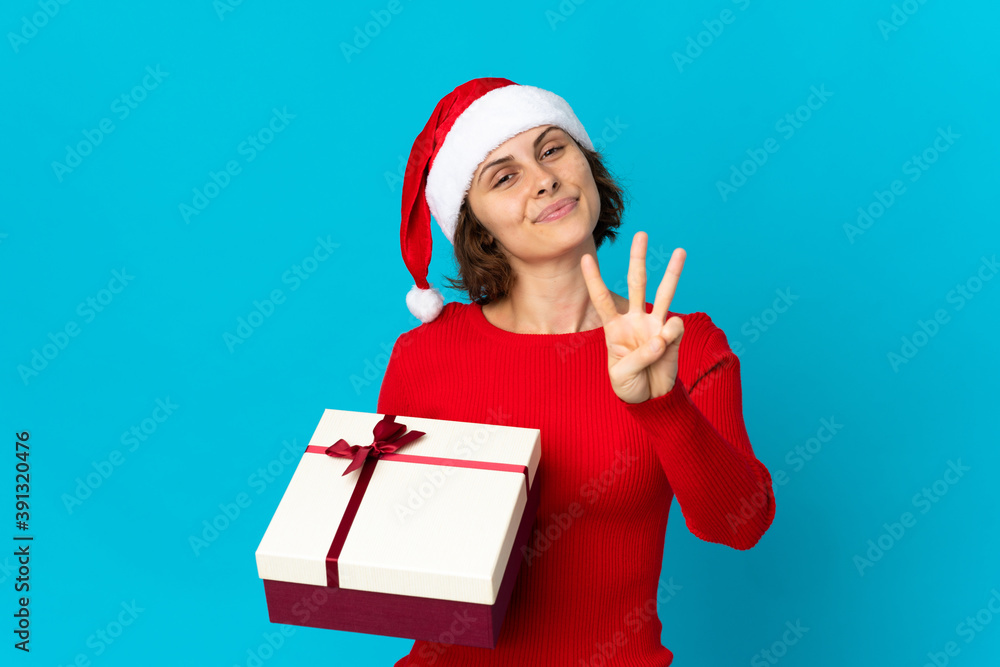 English girl with christmas hat holding a present isolated on blue background happy and counting three with fingers English girl with christmas hat holding a present isolated on blue background happy and counting three with fingers