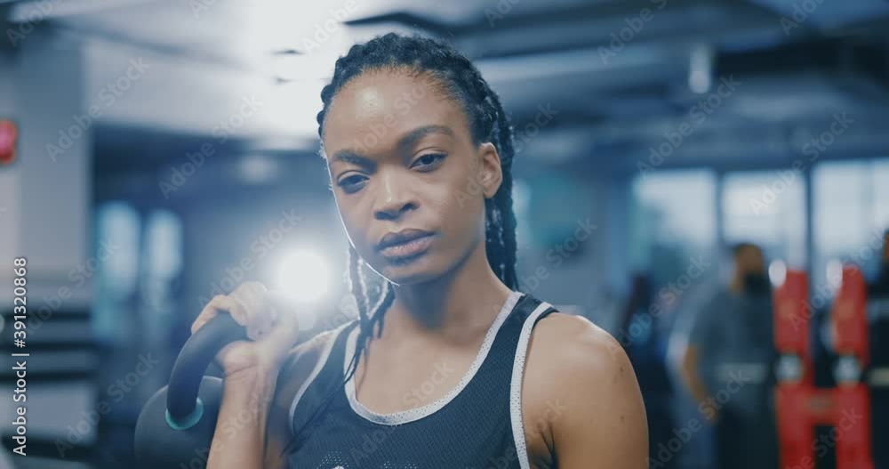 Portrait of powerful african american strong sportswoman with braids holding dumbbells looking confident at camera posing inside gym. Sportswoman. Attractive look. Active lifestyle. Portrait of powerful african american strong sportswoman with braids holding dumbbells looking confident at camera posing inside gym. Sportswoman. Attractive look. Active lifestyle.