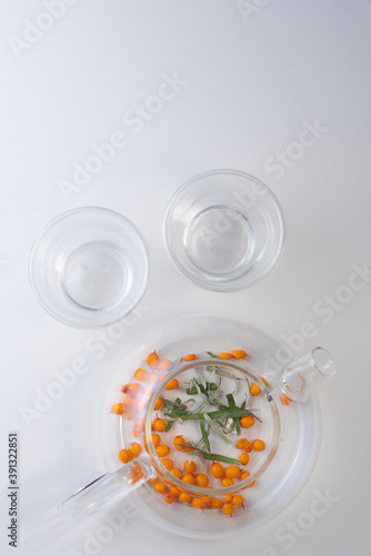 sea buckthorn berries are poured into a transparent teapot on a white table and two transparent mugs next to it. Flat lay