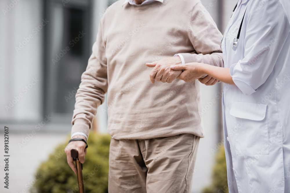 cropped view of social worker supporting aged man strolling with walking stick
