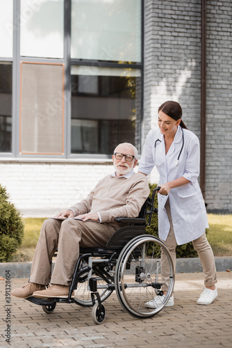 cheerful social worker walking with elderly handicapped man in wheelchair outdoors
