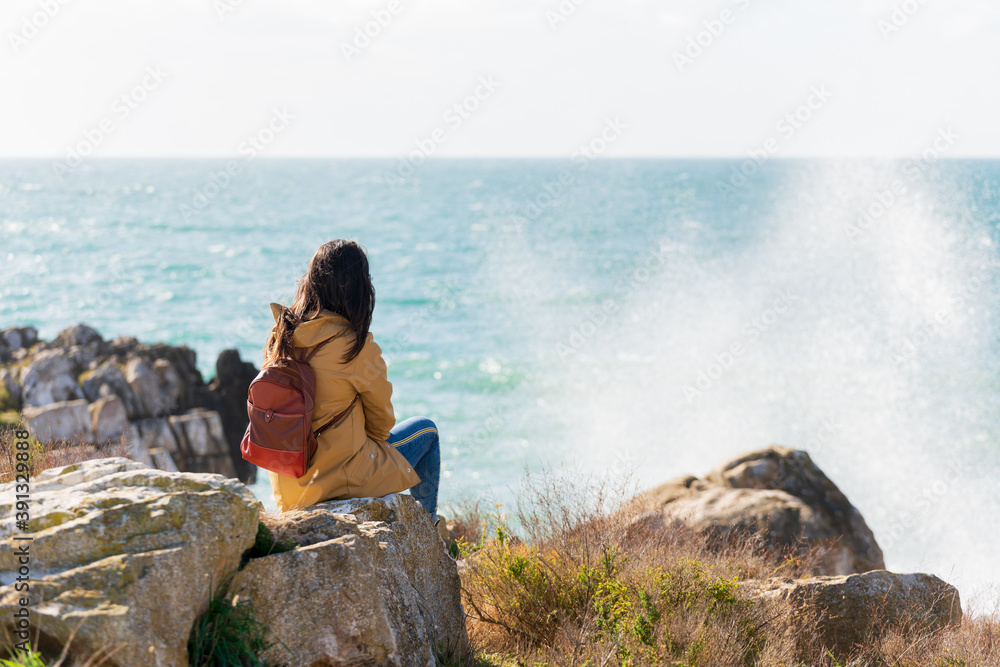 Caucasian traveler woman looking at the waves on the sea with a yellow jacket in Peniche, Portugal