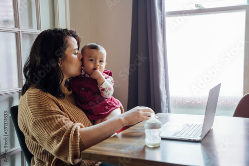 Mom holding baby and working from home on laptop at dining room table 