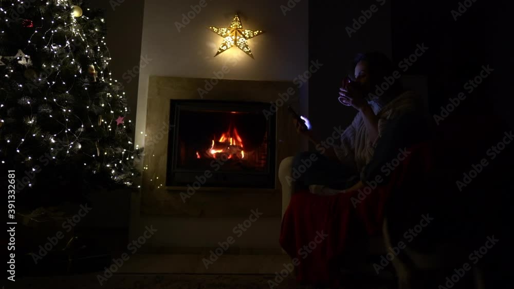 A young woman sits alone near the fireplace and a Christmas tree with a phone in her hands. Social distance. Christmas during lockdown, new year and the covid-19 coronavirus pandemic A young woman sits alone near the fireplace and a Christmas tree with a phone in her hands. Social distance. Christmas during lockdown, new year and the covid-19 coronavirus pandemic