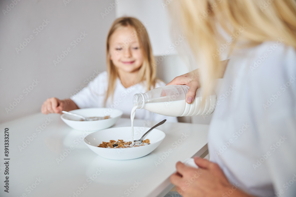 Adult woman pouring milk for their breakfast while she is sitting with little girl in the kitchen home Adult woman pouring milk for their breakfast while she is sitting with little girl in the kitchen home