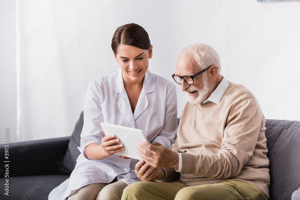 brunette social worker showing digital tablet to smiling aged man at home brunette social worker showing digital tablet to smiling aged man at home