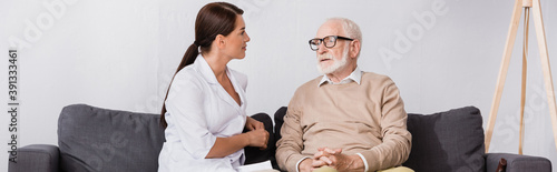 geriatric nurse talking to aged man while sitting on sofa at home, banner